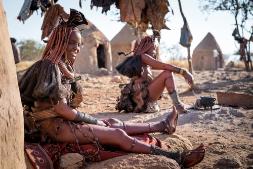 Two women of the Himba people sit in the foreground with traditional Himba huts in the background.