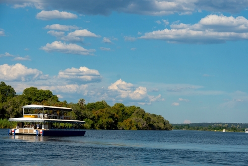 Tourist boat cruising down the middle of the Zambezi River with a tree covered bank in the background.