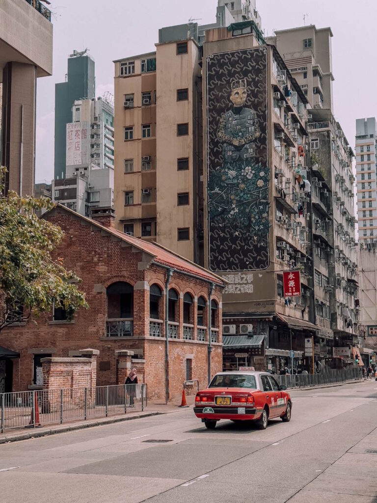 Street view of Yau Ma Tei neighbourhood in Hong Kong, with a red taxi passing an old red brick theatre and more modern buildings with graffiti art on the side. 