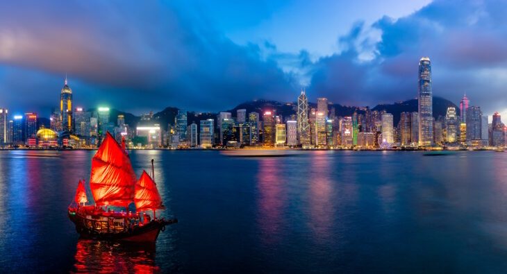 A view of the Hong Kong skyline at twilight. The background is skyscrapers lit up with a boat with red sails on the water in front.