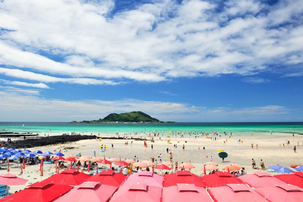 Looking out to sea with an island in the distance, the yellow sand beach full of people enjoying the sunshine, red umbrellas