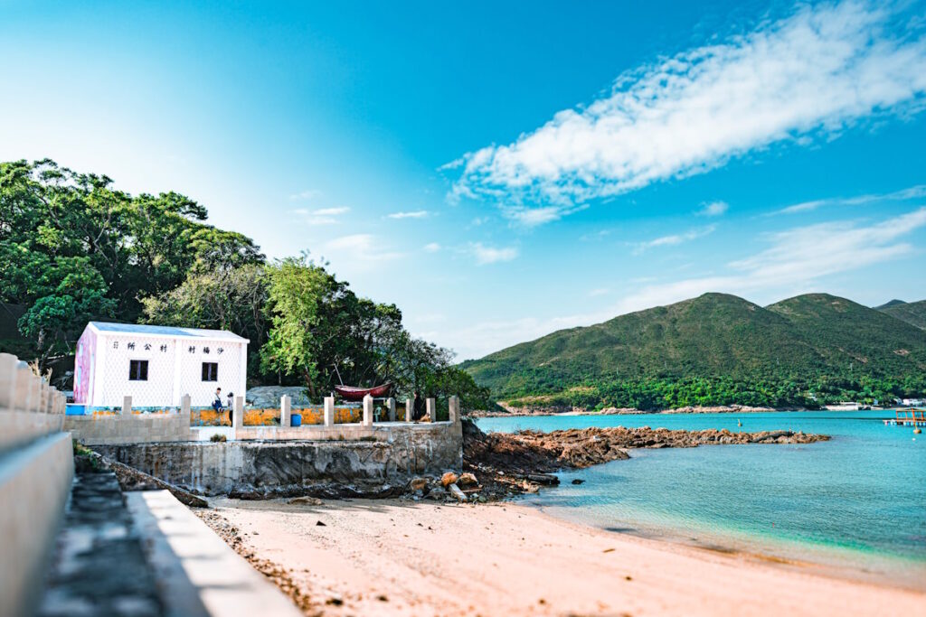 View along a yellow sand beach towards a white building, the blue water of the bay lapping a hilly, greenery covered coastline. 