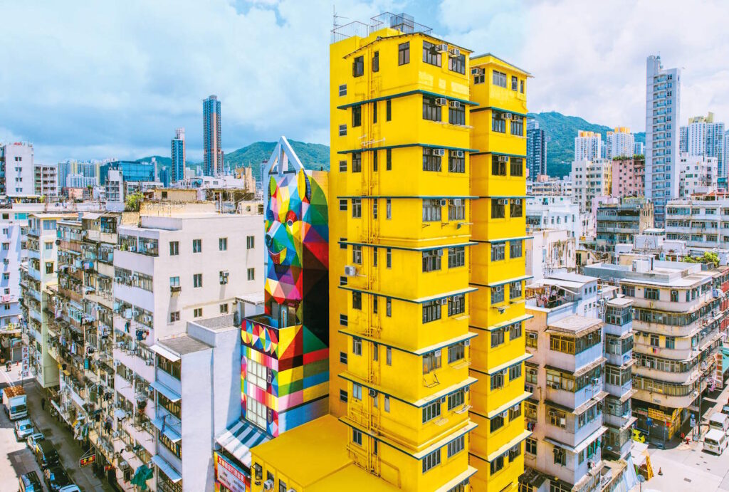 High up view of the tower blocks of Sham Shui Po, centred around a bright yellow building with a colourful piece of artwork featuring a bear on the building next door. 