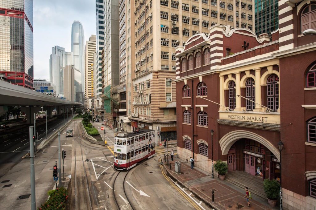 Looking down a street with a tram coming around the corner past an old red market market building, modern skyscrapers in the background. 