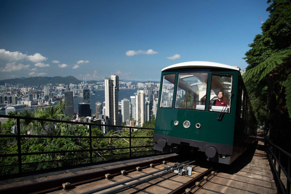 The green Victoria Peak tram running up the track with a view of Hong Kong's skyscrapers in the background under blue skies.