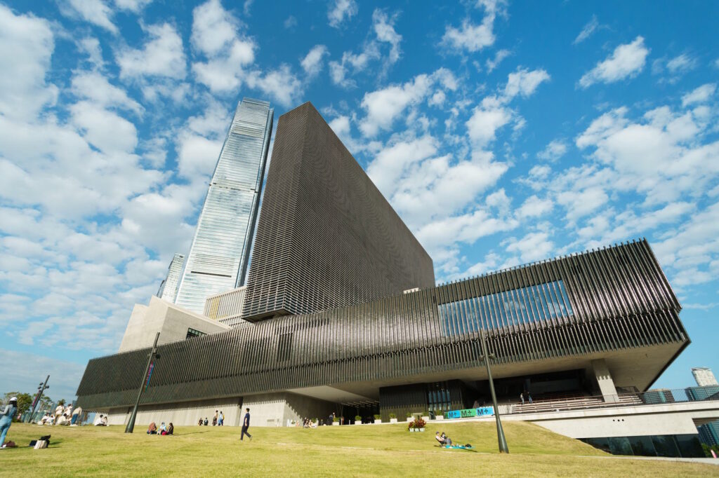 Low angle view of the modern architecture of M+, a contemporary art museum, with grass in the foreground and blue sky behind.