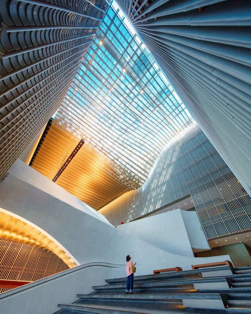 Light shines through the glass ceiling as a women stares up into the airy, open space created by the modern architecture of the Hong Kong Palace Museum