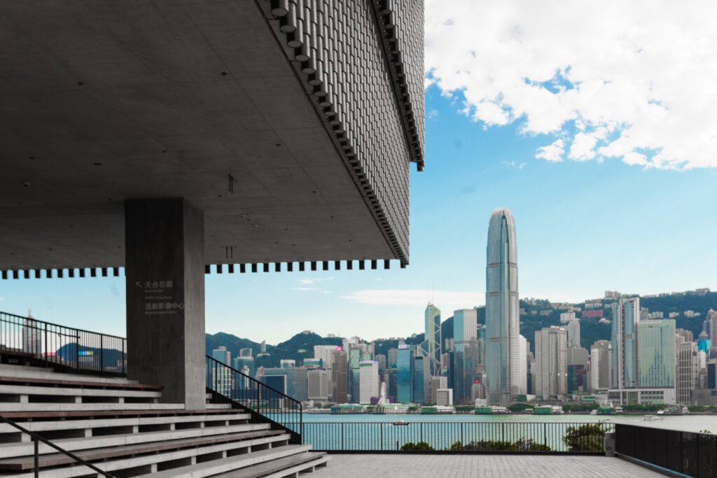 Part of a modern building shows in the top left corner with a view of the Hong Kong skyline over the waters of Victoria Harbour behind.