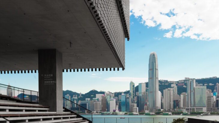 Part of a modern building shows in the top left corner with a view of the Hong Kong skyline over the waters of Victoria Harbour behind.