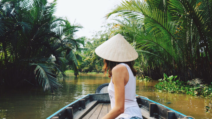 A woman in a conical hat sits in the front of a wooden boat cruising through a palm-lined canal in the Mekong Delta, Vietnam