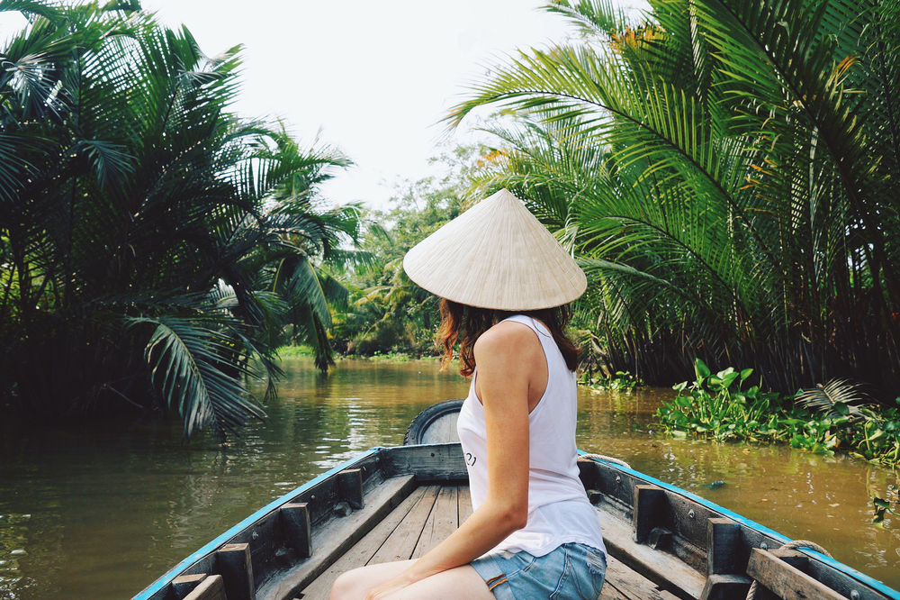 A woman in a conical hat sits in the front of a wooden boat cruising through a palm-lined canal in the Mekong Delta, Vietnam