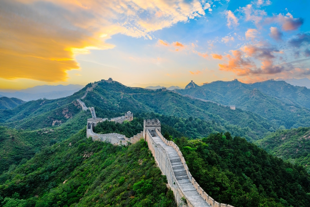 A scenic view of the Great Wall of China winding through lush green mountains under a vibrant sunset sky with orange, pink, and blue hues.