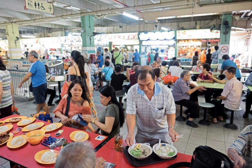 Groups of people sit at tables enjoying food from the many stalls at a Singapore hawker centre.