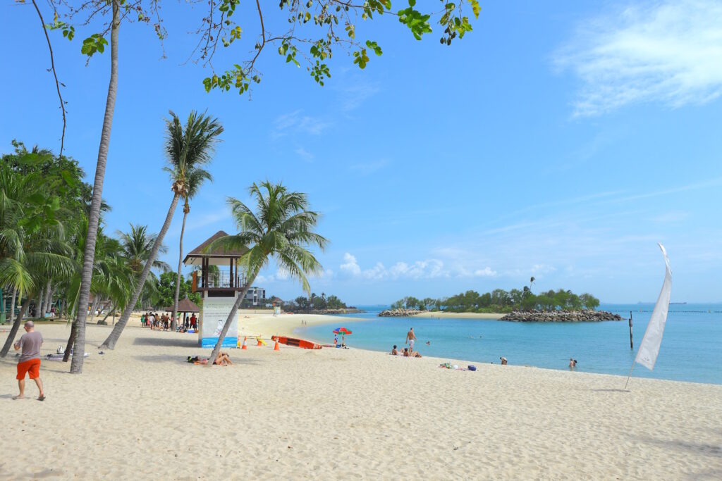 Sunny day at Sentosa Beach, Singapore, featuring soft golden sand, palm trees, a lifeguard tower, and people enjoying the clear blue waters.