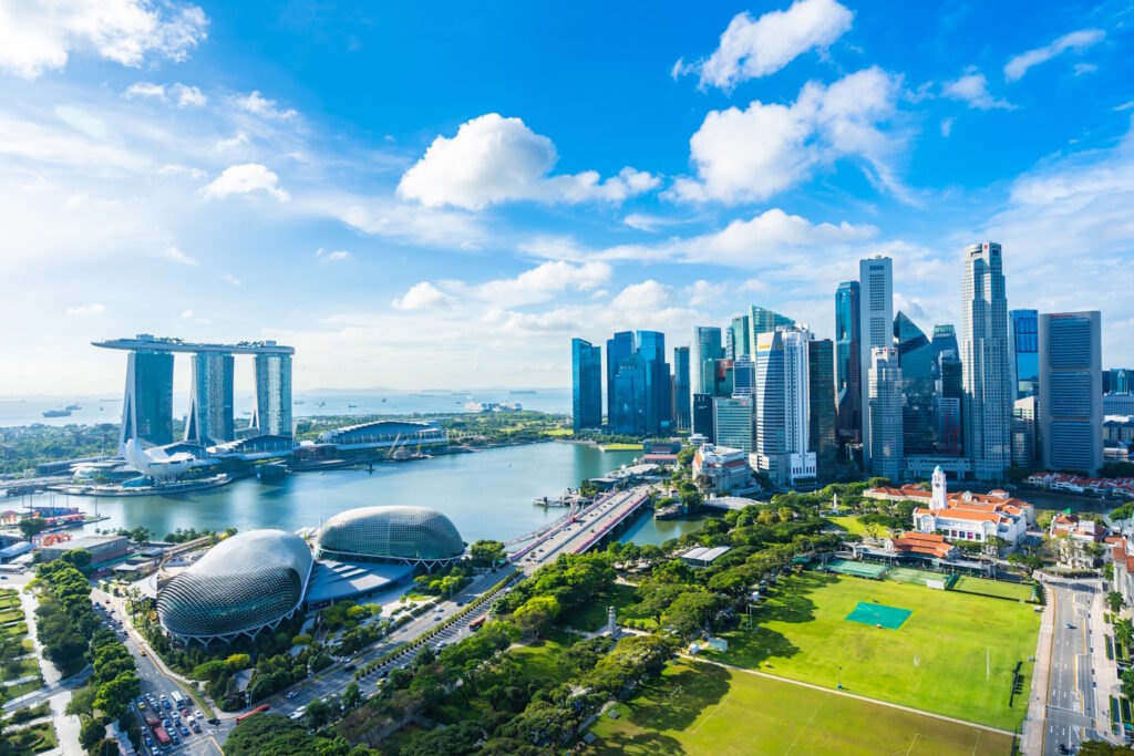 Aerial view of Singapore’s Marina Bay area, featuring Marina Bay Sands, Esplanade Theatres, the central business district skyline, and lush green spaces under a bright blue sky.