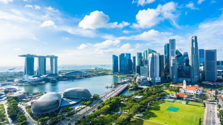 Aerial view of Singapore’s Marina Bay area, featuring Marina Bay Sands, Esplanade Theatres, the central business district skyline, and lush green spaces under a bright blue sky.